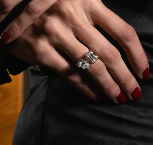 A detailed shot of a hand wearing two lab-grown diamond rings—a radiant-cut and a pear-cut—set against a sleek black dress. The red nails and soft lighting highlight the brilliance of the jewelry.