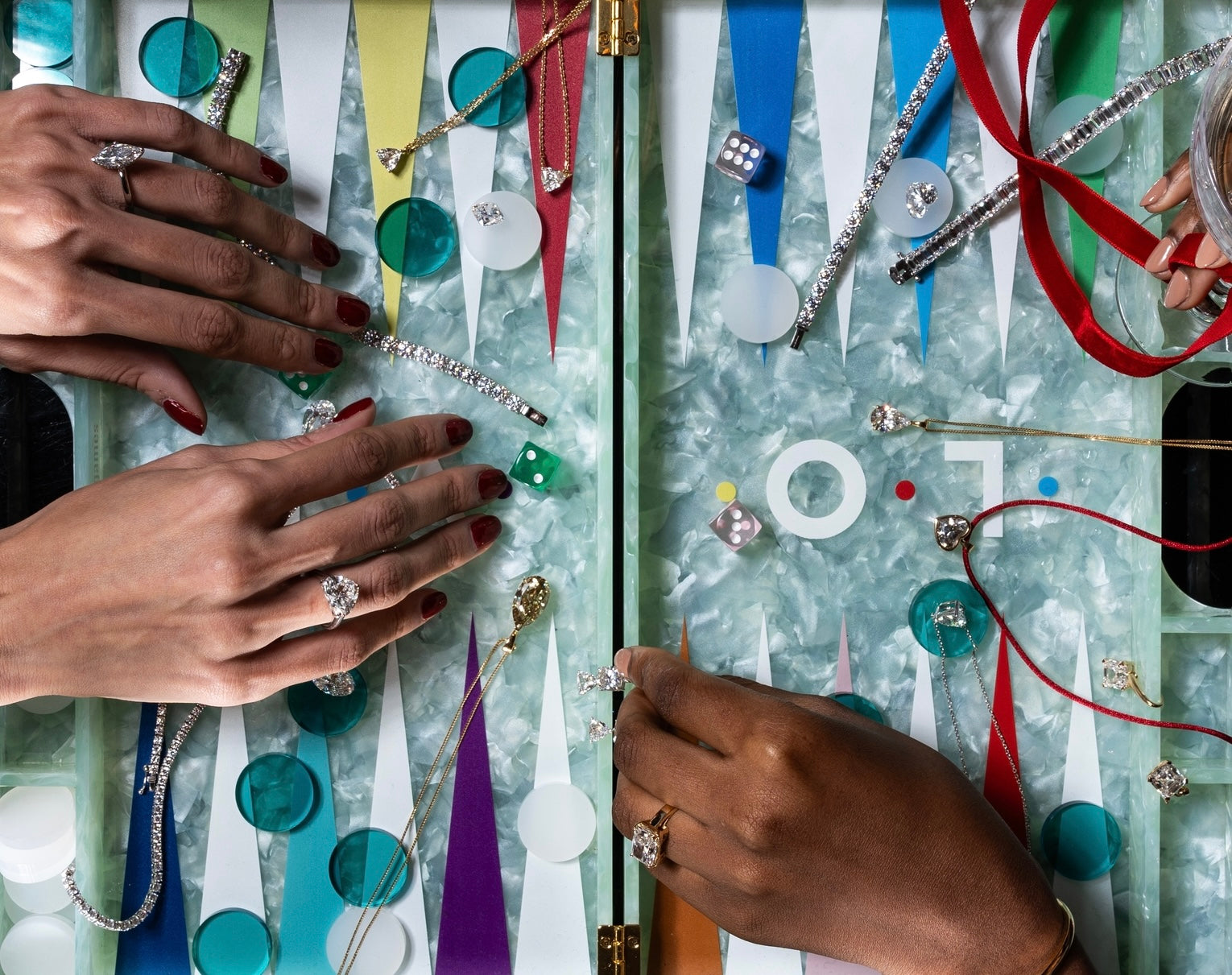 An overhead view of a colorful backgammon board adorned with lab-grown diamond jewelry, including rings, bracelets, and necklaces. Hands with polished nails and elegant diamond rings are positioned around the board, adding a luxurious touch to the playful scene.