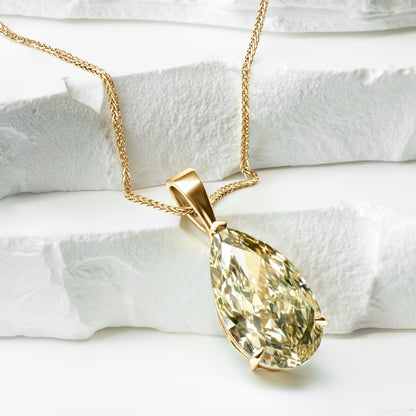 A close-up of a pear-shaped lab-grown yellow diamond pendant, elegantly set in a gold setting and hanging from a delicate gold chain. The pendant is displayed on a textured white surface, highlighting its brilliance and craftsmanship.