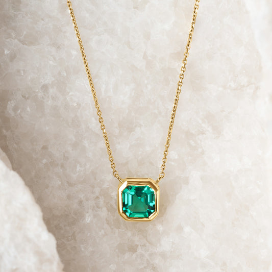  A refined lab-grown emerald asscher-cut green gemstone pendant in a gold bezel setting, elegantly suspended on a delicate gold chain. The piece is displayed on a textured white crystalline background, emphasizing its vibrant hue and modern design.