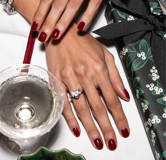 Close-up of hands with polished red nails, one adorned with a stunning heart-shaped lab-grown diamond ring. The scene is styled on a holiday-themed table featuring a wrapped gift, a glass of sparkling liquid, and a decorative ribbon.